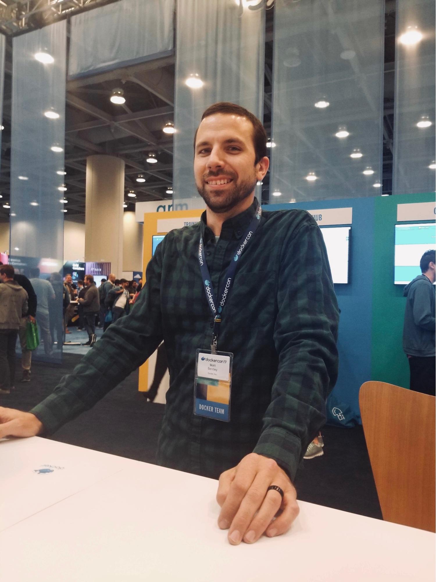 Matt Betley standing behind a booth counter at DockerCon 2019 with hands on counter wearing "Docker Team" badge