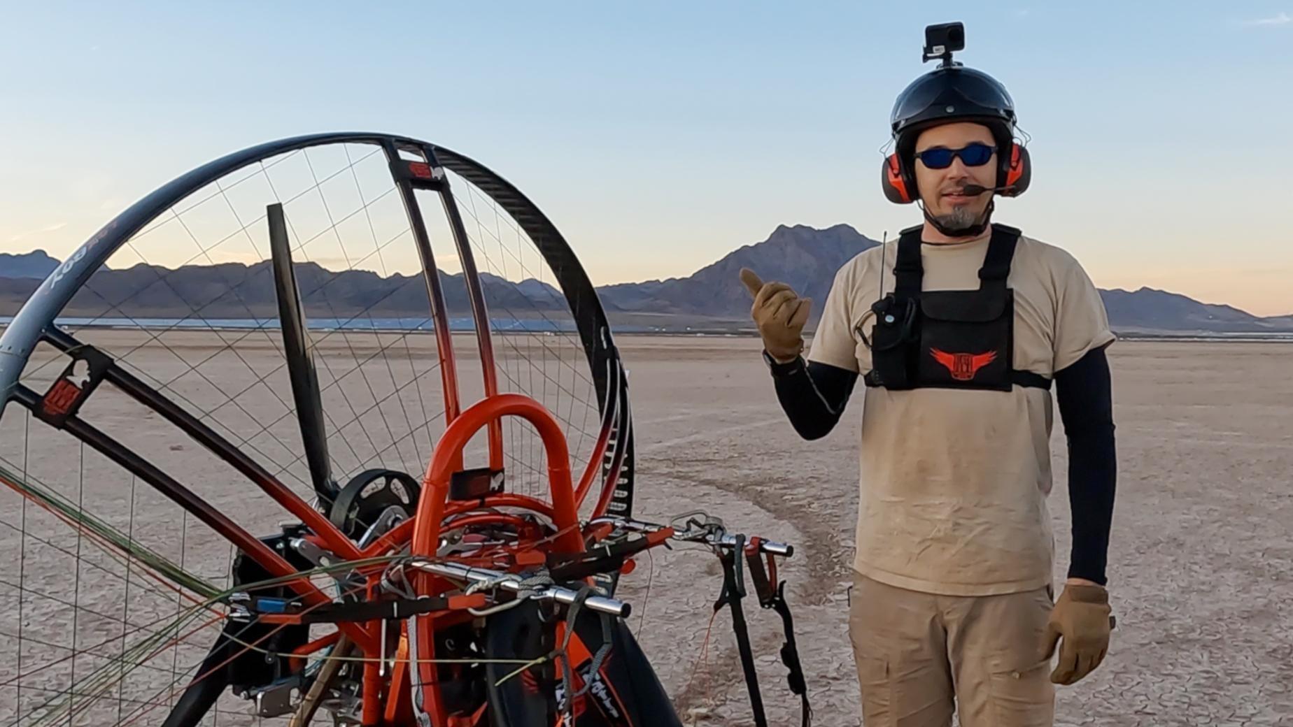 Adrian Goins in helmet and various bits of camera equipment standing next to an orange and black paramotor quad aircraft