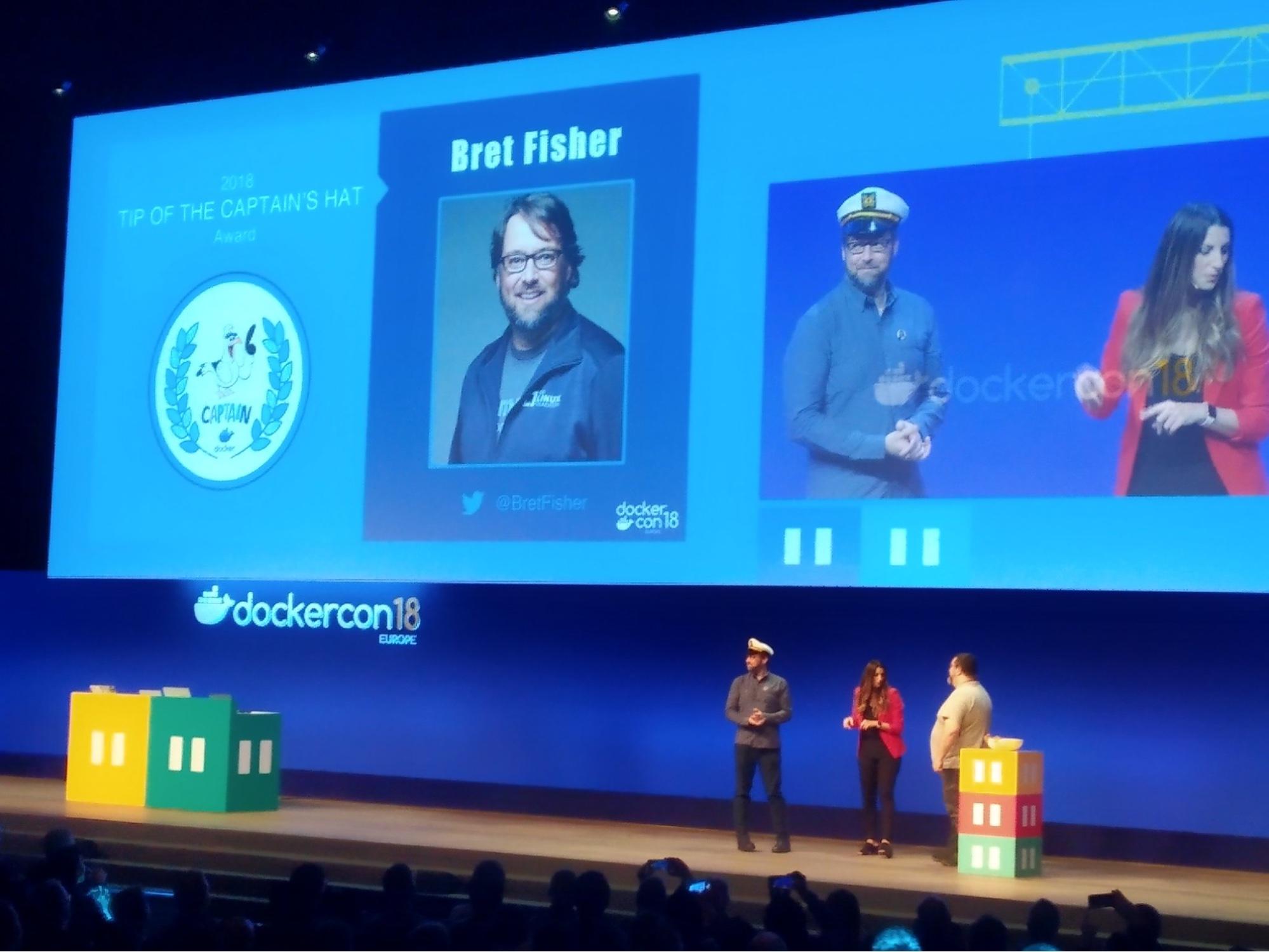 DockerCon 18 Europe keynote stage with Jenny Burcio and Mano Marks presenting Bret Fisher with the "Top of the Captains Hat" award. Bret is wearing a white boating captains hat.